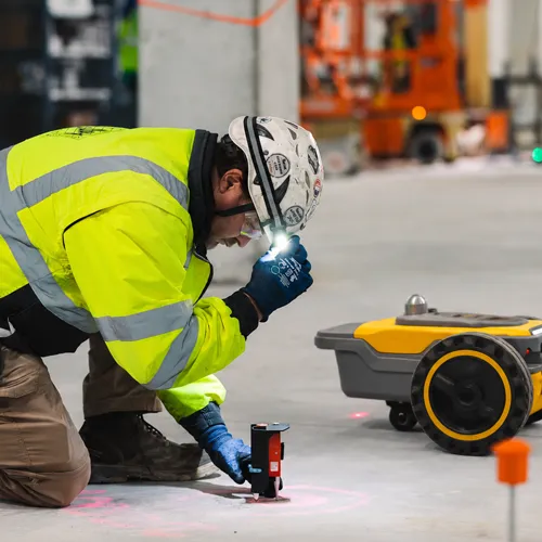 Technicien à genoux sur la dalle posant un repère au sol, le robot traceur à côté de lui