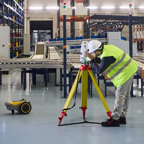 Technicien au casque blanc réglant un instrument de mesure sur trépied jaune, avec le robot traceur sur la dalle
